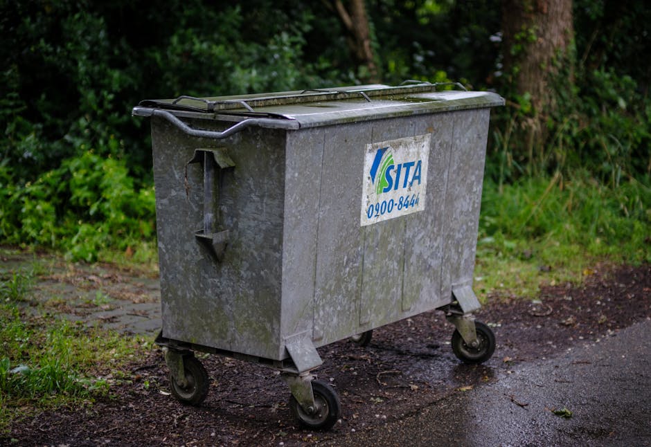 A grey, weathered wheelie bin with a partially visible sticker displaying the SITA logo and contact number, situated outdoors on a damp, uneven pavement near a grassy area with dense green foliage and